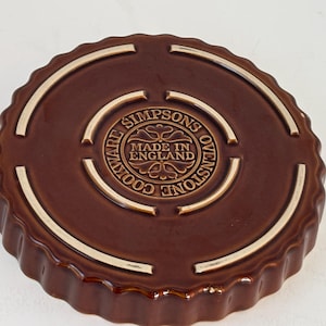 Può includere: Pirofila rotonda marrone in gres con bordo scanalato. La base presenta un emblema centrale con il testo "SIMPSONS OVENSTONE COOKWARE MADE IN ENGLAND". La pirofila ha una finitura lucida ed è progettata per l'uso in forno.
