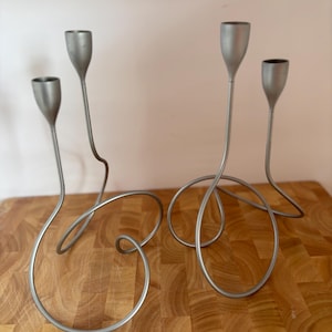 Può includere: Due set di candelabri in metallo color argento. Ogni candelabro ha un gambo sottile e curvo che forma un anello alla base. La parte superiore di ogni candelabro ha un supporto a forma di coppa per una candela. I candelabri sono su una superficie di legno.