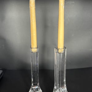 May include: Two clear crystal candle holders with a faceted base and a tall, slender body. Each holder contains a wrapped, light-yellow candle. The candle holders have a logo sticker on the base.