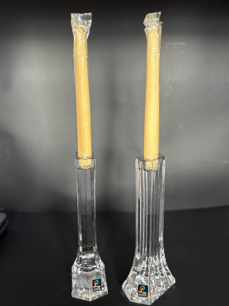 May include: Two clear crystal candle holders with a faceted base and a tall, slender body. Each holder contains a wrapped, light-yellow candle. The candle holders have a logo sticker on the base.