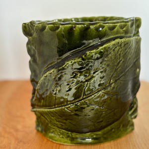 Peut inclure: Un vase en céramique vert foncé avec une finition brillante. Le vase présente un motif de feuilles détaillé autour du corps cylindrique. Le bord a un bord festonné texturé, ajoutant à son esthétique unique.