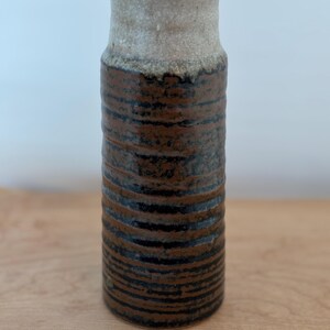 Könnte beinhalten: Eine zylindrische Keramikvase mit einem strukturierten, geriffelten Design. Die Vase hat ein braunes und schwarzes Streifenmuster im unteren Bereich, das in eine hellbeige Farbe übergeht. Die Vase ist etwa 25 cm hoch.