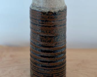Könnte beinhalten: Eine zylindrische Keramikvase mit einem strukturierten, geriffelten Design. Die Vase hat ein braunes und schwarzes Streifenmuster im unteren Bereich, das in eine hellbeige Farbe übergeht. Die Vase ist etwa 25 cm hoch.