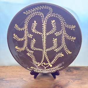 Puede incluir: Un plato redondo de cerámica con un esmalte de color ciruela intenso que presenta un diseño botánico pintado a mano en amarillo claro, con un árbol central de ramas simétricas y frondosas y un pequeño corazón en la base, con borde ondulado.