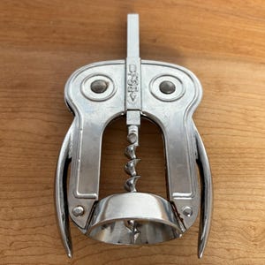 Könnte beinhalten: Ein silberner Korkenzieher mit einem einzigartigen Design. Er hat einen Griff, eine Wendel und einen glockenförmigen Sockel. Das Design umfasst zwei kreisförmige, augenähnliche Merkmale. Der Korkenzieher aus Metall ist zum Entfernen von Korken aus Weinflaschen bestimmt.