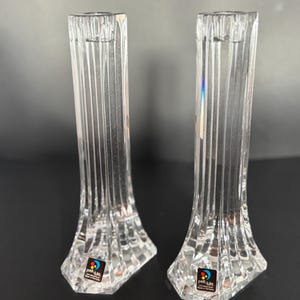 May include: Two clear, crystal candle holders with vertical ribbed detailing. The candle holders have a flared base and a narrow top designed to hold a candle. Each holder has a colorful sticker on the base.