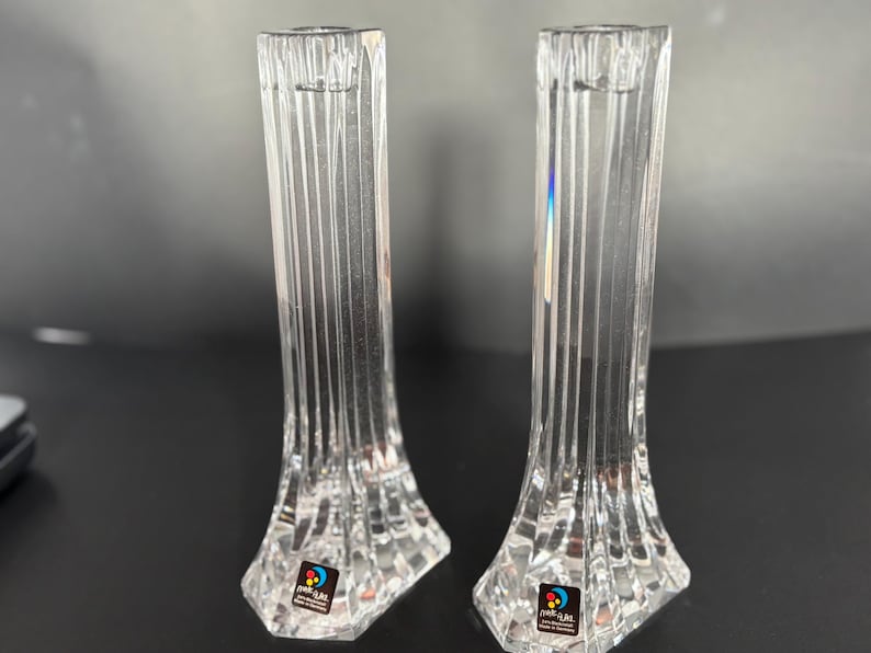 May include: Two clear, crystal candle holders with vertical ribbed detailing. The candle holders have a flared base and a narrow top designed to hold a candle. Each holder has a colorful sticker on the base.