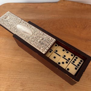 Peut inclure: Un jeu de dominos en bois avec un couvercle décoratif en argent. Les dominos sont de couleur ivoire avec des points noirs. La boîte en bois est marron foncé.