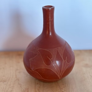 May include: A terracotta-coloured ceramic vase with a rounded body and a slender neck. The vase features an etched leaf design around the body. The vase is displayed on a wooden surface.