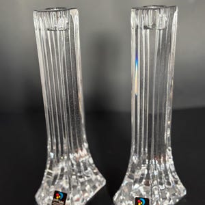 May include: Two clear crystal candle holders. Each holder has a rectangular shape with vertical grooves and a faceted base. The candle holders are labeled with a colorful sticker. The background is dark.