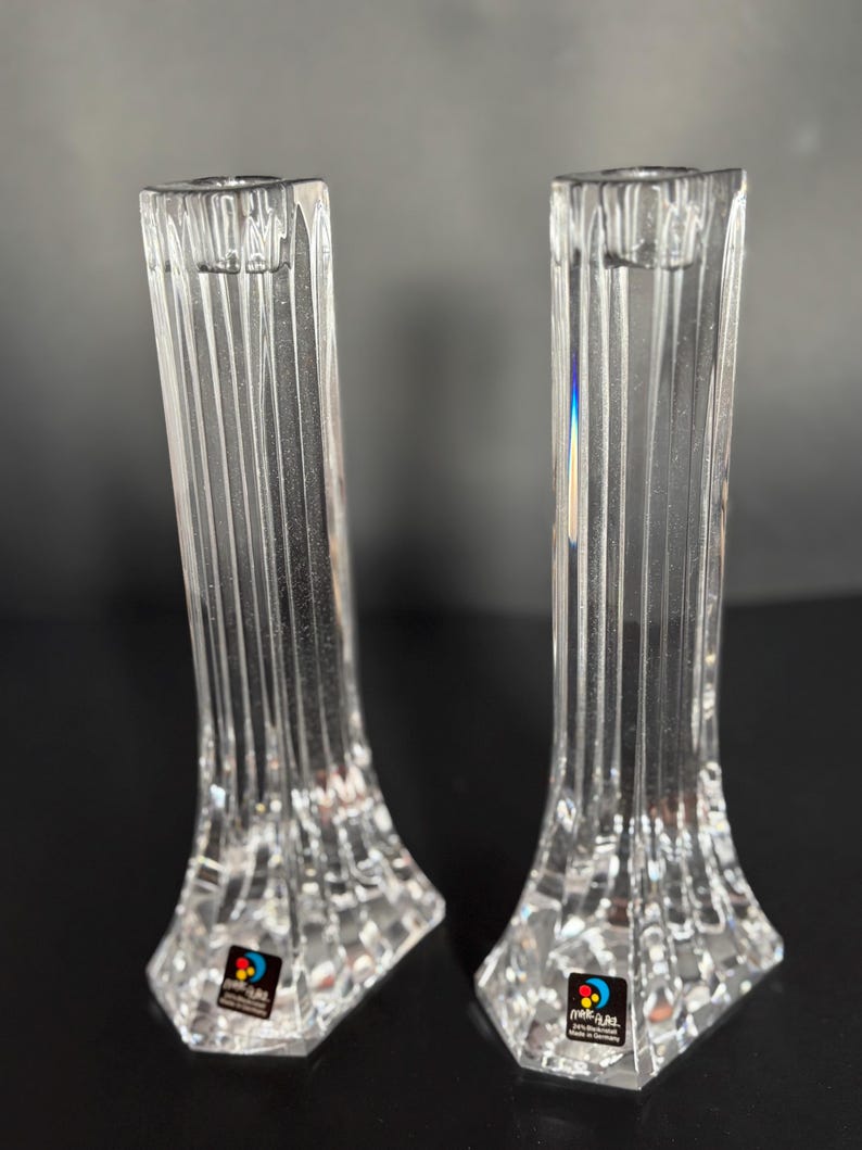 May include: Two clear crystal candle holders. Each holder has a rectangular shape with vertical grooves and a faceted base. The candle holders are labeled with a colorful sticker. The background is dark.