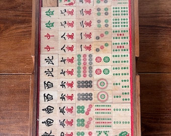 Puede incluir: Una caja rectangular de madera que contiene un juego completo de fichas de Mahjong dispuestas en filas ordenadas. Las fichas de madera clara presentan varios caracteres chinos tradicionales, patrones circulares y símbolos de bambú en tinta roja, verde y negra.