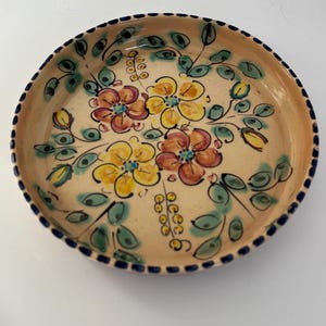 Op de afbeelding: Een ronde, handbeschilderde keramische plaat met een beige achtergrond. De plaat heeft een bloemenontwerp met gele, oranje en roze bloemen, groene bladeren en een donkerblauwe rand. De plaat heeft een diameter van ongeveer 25 cm.