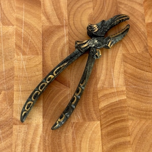 May include: Antique brass nutcracker with a detailed design. The nutcracker features a dark finish with gold accents. The handles have a curved shape with decorative elements. The jaws are open, ready for use. The nutcracker is on a wooden surface.