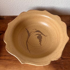 Könnte beinhalten: Eine beigefarbene, gesprenkelte Keramikschale mit gewelltem Rand und einer braunen, handgemalten botanischen Illustration eines Weizenhalms in der Mitte. Die Schale hat eine matte Oberfläche und ein breites, flaches Becken mit gewelltem Rand.