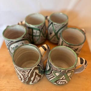 Peut inclure: Six tasses en céramique faites à la main avec des motifs uniques. Chaque tasse présente une base crème avec des motifs abstraits noirs et verts et une anse assortie. Les bords sont peints en vert, et les tasses sont disposées sur une surface en bois.