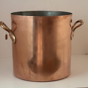 Op de afbeelding: Een koperen pot met twee messing handvatten. De pot heeft een cilindrische vorm en een glanzend oppervlak. De handvatten zijn met klinknagels bevestigd. Dit keukengerei is geschikt om te koken.