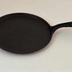Puede incluir: Una plancha de hierro fundido negro con un solo mango. La sartén es redonda con una superficie de cocción plana y un borde elevado. El mango tiene un lazo para colgar. Ideal para cocinar panqueques o tortillas.