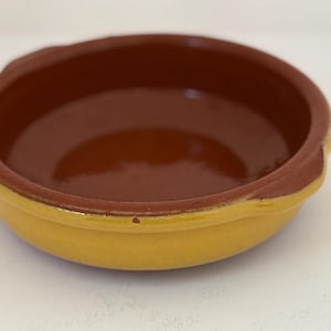 Op de afbeelding: Een kleine, ronde keramische kom met een gele buitenkant en een bruine binnenkant. De rand is donkerbruin. Deze rustieke kom is geschikt voor het serveren van kleine porties of als decoratief stuk. Diameter ongeveer 12 cm.