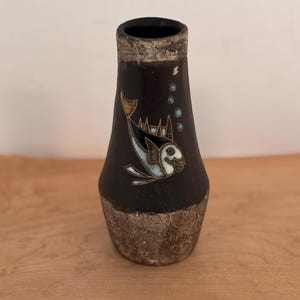 Può includere: Un vaso decorativo nero con un disegno di pesce. Il vaso ha una base e un bordo strutturati, marrone chiaro. Il pesce è bianco, nero e oro, con bolle intorno. Il vaso è su una superficie di legno.