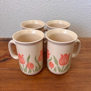 Op de afbeelding: Vier beige keramische mokken met een tulpontwerp. De mokken hebben een crèmekleurige basis met rode tulpenillustraties en groene bladeren. De mokken zijn gerangschikt op een houten oppervlak.