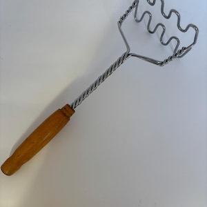 Könnte beinhalten: Ein Kartoffelstampfer mit Holzgriff und einem gedrehten Metallschaft. Der Stampferkopf hat ein wellenförmiges Design. Der Griff ist hellbraun.