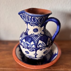 Può includere: Un set composto da brocca e bacinella in ceramica con un motivo floreale dipinto a mano in blu e bianco. La ceramica presenta un bordo e un interno color terracotta, con intricati disegni geometrici a griglia e motivi floreali stilizzati.