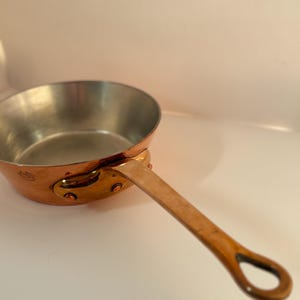 Può includere: Una piccola casseruola di rame con un lungo manico curvo. L'interno della pentola è color argento, in contrasto con l'esterno in rame. Il manico è fissato con rivetti. Un classico articolo per la cucina.