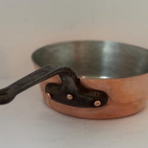 May include: A copper saucepan with a dark, riveted handle. The interior is silver. The pan is designed for cooking and has a classic, elegant look. The handle is attached with copper rivets.