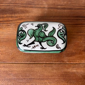 May include: A rectangular ceramic box with a white base and a hand-painted green bird illustration on the lid. The bird is depicted in profile with stylised feathers and black outlines. Decorative green scrollwork accents the sides of the lid.