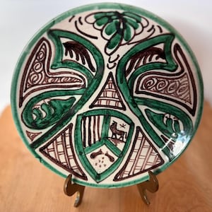 Può includere: Piatto decorativo in ceramica con bordo verde e intricati disegni marroni e verdi. Il piatto presenta un emblema centrale con una figura animale stilizzata e motivi geometrici. È esposto su un supporto color oro.