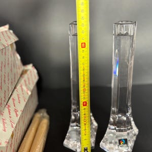 May include: Two clear crystal candle holders, approximately 10 inches tall, with a faceted base and a square top. A yellow measuring tape is placed next to the candle holders. Boxes and candles are in the background.
