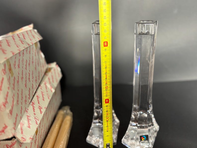 May include: Two clear crystal candle holders, approximately 10 inches tall, with a faceted base and a square top. A yellow measuring tape is placed next to the candle holders. Boxes and candles are in the background.