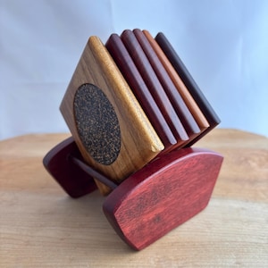 Op de afbeelding: Een set houten onderzetters in een houder. De onderzetters zijn vierkant met verschillende houttinten, waaronder lichtbruin, donkerbruin en roodbruin. De houder is dieprood van kleur en de onderzetters hebben een kurkdetail.
