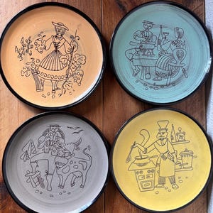 Op de afbeelding: Een set van vier ronde keramische borden met zwarte randen. Elk bord heeft een andere achtergrondkleur—oranje, lichtblauw, grijs en geel—en is versierd met een zwarte lijntekening die verschillende scènes met mensen en dieren toont.