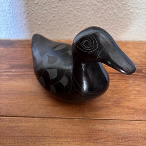 May include: A smooth, black, carved stone duck figurine featuring etched circular patterns around the eye and a repeating scale-like design across the body. The sculpture has a polished finish and a rounded, stylized form with a distinct beak.
