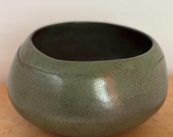May include: A handcrafted, olive green ceramic bowl with a textured surface. The bowl has a rounded shape with a dark interior and a subtle, curved design element near the top. The bowl is a decorative piece.