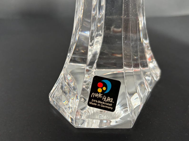 May include: Clear crystal decorative object with a faceted design. The base is hexagonal, and the object has a flared shape. A sticker on the base reads "MARC AUREL 24% Bleikristall Made in Germany."