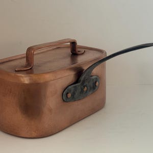 Può includere: Una pentola rettangolare in rame con un manico sul coperchio e un lungo manico curvo attaccato al lato. La pentola ha una superficie lucida e un attacco del manico rivettato scuro.