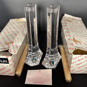 May include: Two clear crystal candle holders with a faceted design, each with a matching box and a candle. The boxes have text and are labeled "Marc Aurel". The candle holders are approximately 10 inches tall.