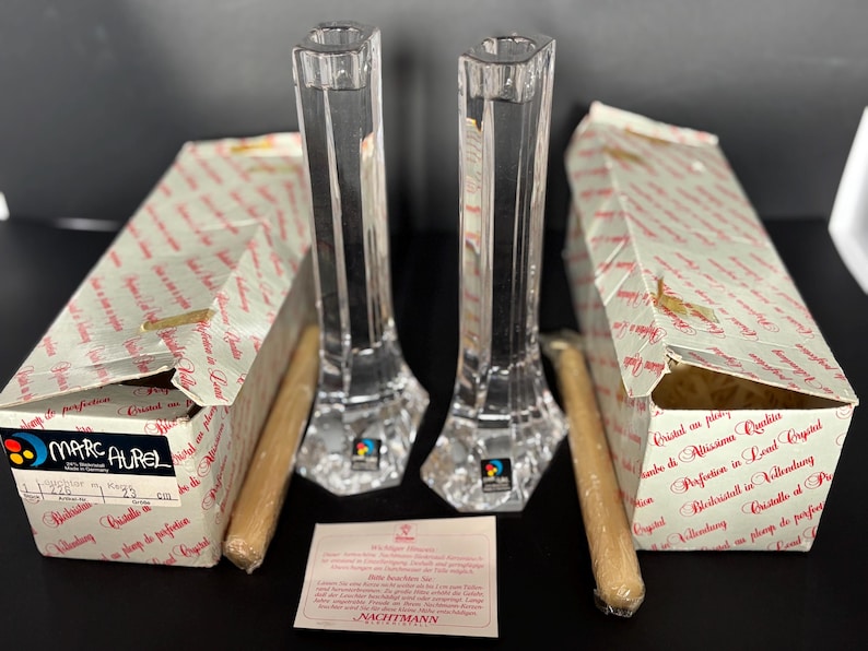 May include: Two clear crystal candle holders with a faceted design, each with a matching box and a candle. The boxes have text and are labeled "Marc Aurel". The candle holders are approximately 10 inches tall.