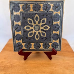May include: Square ceramic tile with a floral design. The tile has a navy blue centre with a cream-coloured flower and a border of navy blue floral accents on a cream background. The tile is displayed on a small, red wooden easel.