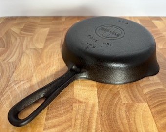 May include: A black Griswold No. 3 cast iron skillet with a long handle. The skillet has the Griswold logo and the words "ERIE PA." and "709 I" embossed on the bottom. The skillet is on a wooden cutting board.