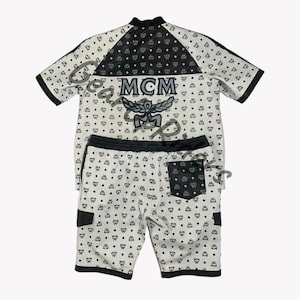 Puede incluir: Un conjunto de dos piezas a juego con una camisa de manga corta y pantalones cortos. El conjunto es principalmente blanco con un patrón negro repetido. La camisa tiene un cuello y un panel de hombro negros, con el logotipo "MCM" y un emblema alado. Los pantalones cortos tienen una cintura y un bolsillo negros.