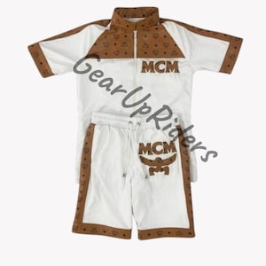 Puede incluir: Conjunto deportivo blanco con pantalones cortos y chaqueta de manga corta con ribetes y detalles marrones. La chaqueta tiene cremallera y las letras "MCM" en la parte delantera. Los pantalones cortos también tienen el logo "MCM".