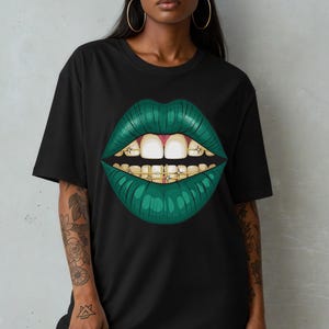 Może przedstawiać: Czarny t-shirt z grafiką zielonych ust i złotym aparatem ortodontycznym. Usta mają fakturę, a zęby są białe. Koszulka ma swobodny, obszerny krój.