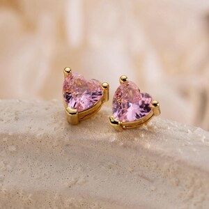 Könnte beinhalten: Ein Paar Ohrstecker mit herzförmigen rosa Edelsteinen in goldfarbenem Metall. Jeder Ohrring hat eine facettierte Oberfläche, die das Licht reflektiert, mit einer spitz zulaufenden Basis und einer abgerundeten Oberseite in einer Metallfassung.