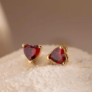 May include: A pair of heart-shaped stud earrings. Each earring has a vibrant red gemstone set in gold-coloured metal. The earrings are displayed on a textured, off-white surface, with a blurred background.