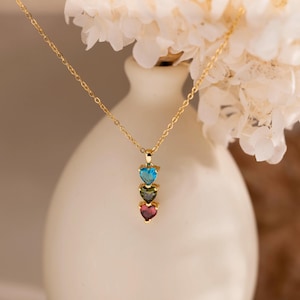 Könnte beinhalten: Eine goldfarbene Halskette mit einem Anhänger aus drei herzförmigen Edelsteinen in Blau, Grün und Rot. Die Halskette wird vor einem verschwommenen Hintergrund mit weißen Blumen präsentiert.