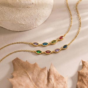 May include: Two gold necklaces with colorful gemstone accents. Each necklace features a delicate chain and a series of oval-shaped gemstones in various colors, including red, green, blue, and purple. The necklaces are displayed on a neutral background.